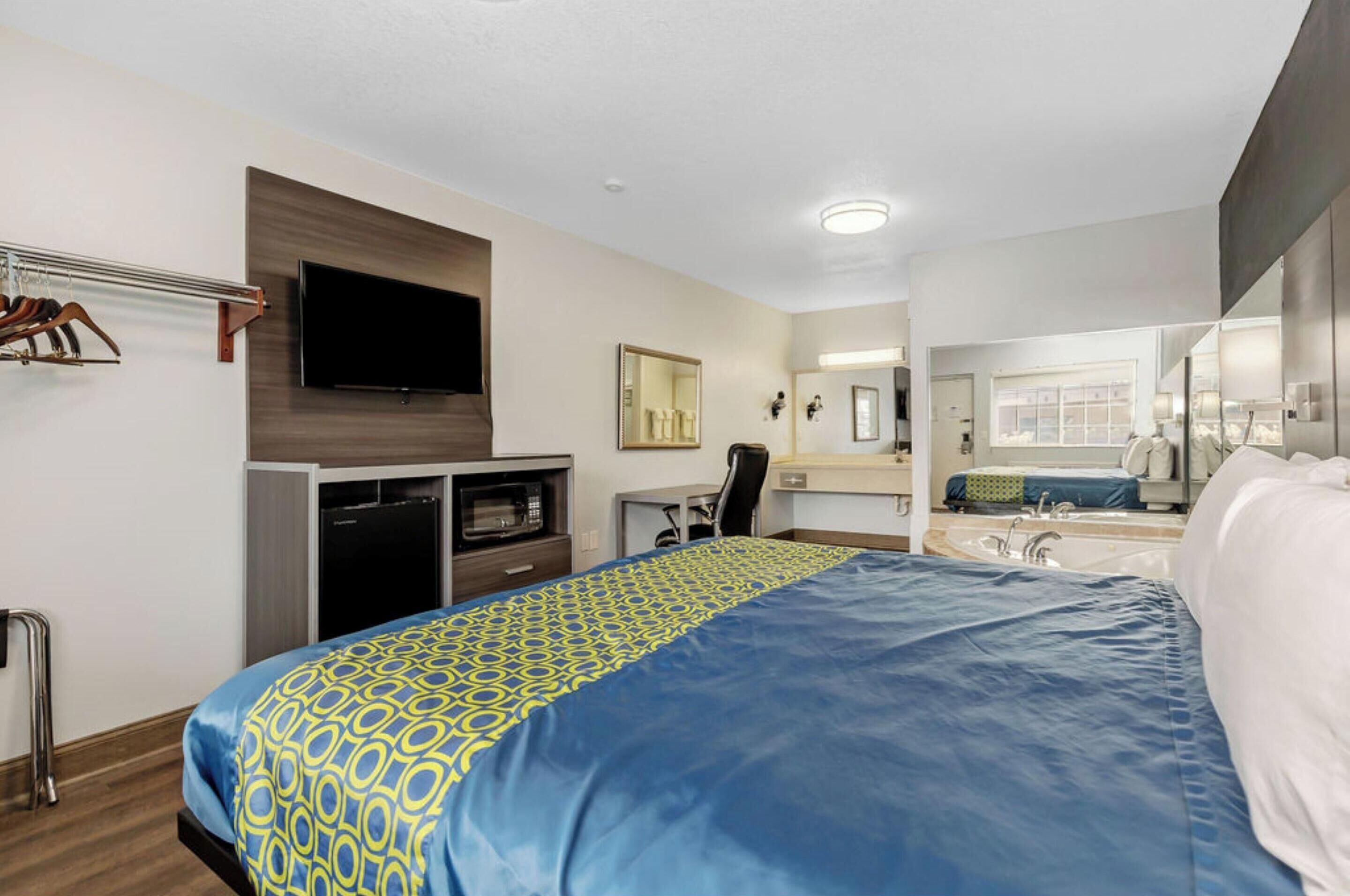 A hotel room with a bed, a desk, a television, a mirror, and a bathroom with a sink and mirror.