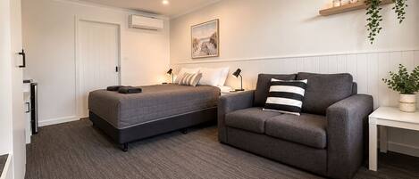 A bedroom with a bed, a sofa, a bedside table, a lamp, a wall-mounted shelf with plants, and a painting.