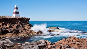 A rocky coastline with a lighthouse on a cliff, waves crashing against the rocks, and a clear blue sky. - Robetown Motel (Robe)