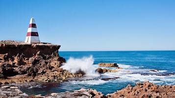 Costa rocosa con un faro en un acantilado, olas rompiendo contra las rocas y un cielo azul despejado.