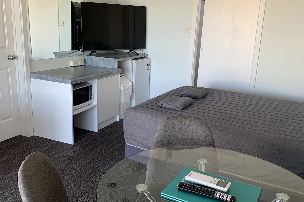 A compact hotel room with a bed, a small desk with a television, a microwave, and a glass table with a remote control.