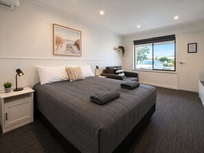 A bedroom with a large bed, bedside tables, a desk, a chair, a window with a view, and a painting on the wall. - Robetown Motel (Robe)