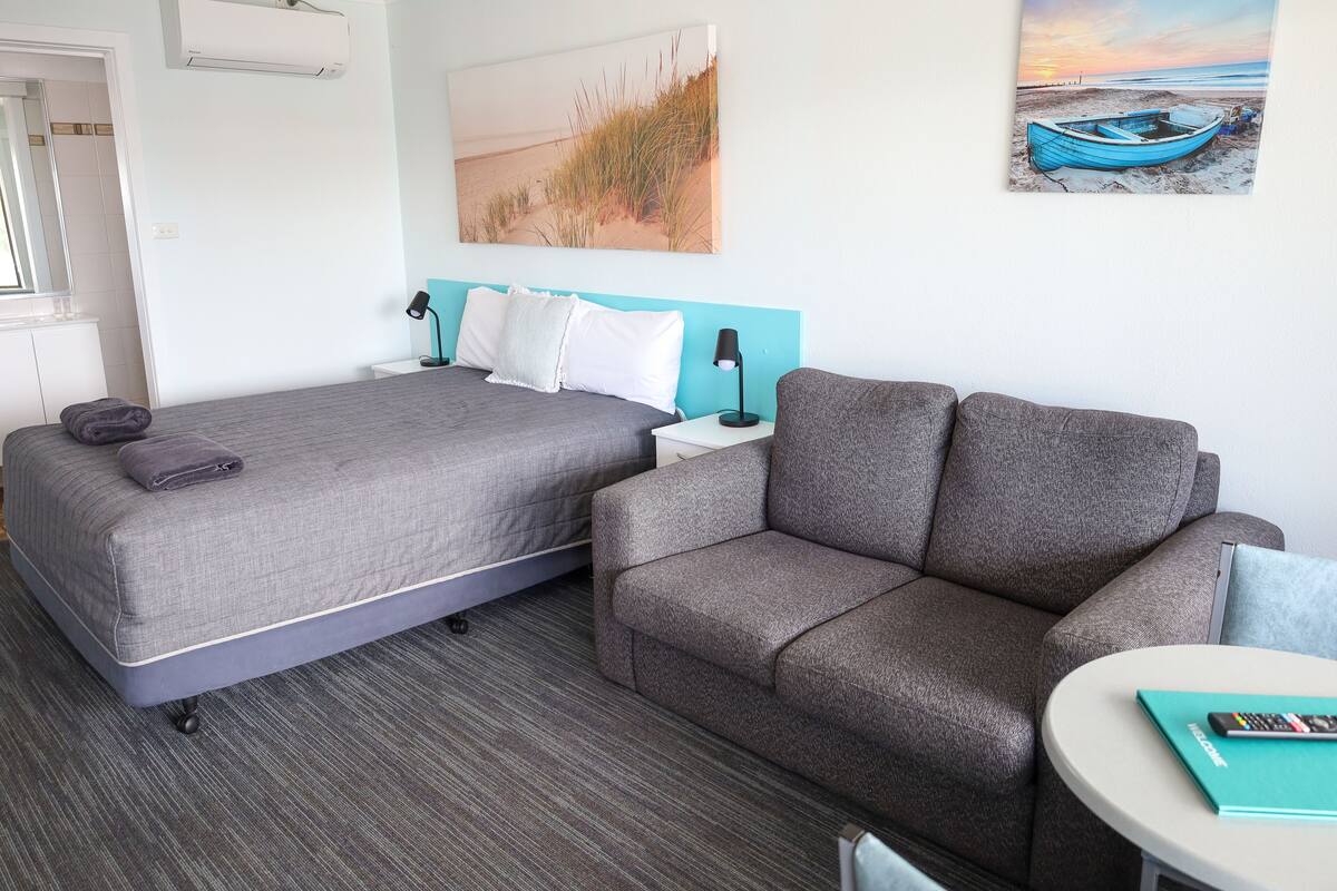 A hotel room with a bed, a sofa, a small table, and two paintings on the wall.