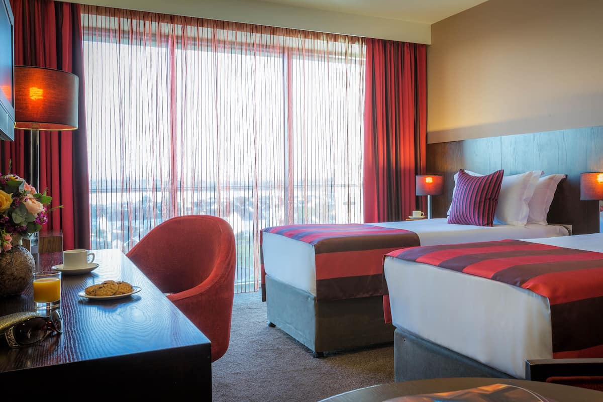 A hotel room with two beds, a desk with a chair, a coffee cup, and a vase with flowers.