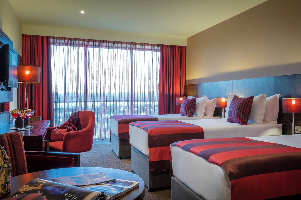 A hotel room with two beds, a desk, a chair, and a view of the city through large windows.