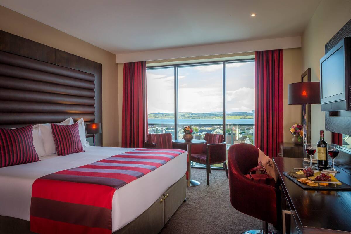 A hotel room with a large bed, a desk with a chair, a TV, and a view of the city.