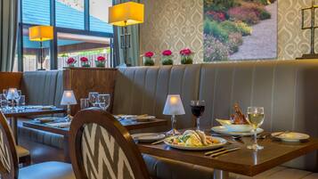 A restaurant with a booth seating area, tables with wine glasses and plates, and a decorative wall with a garden view.