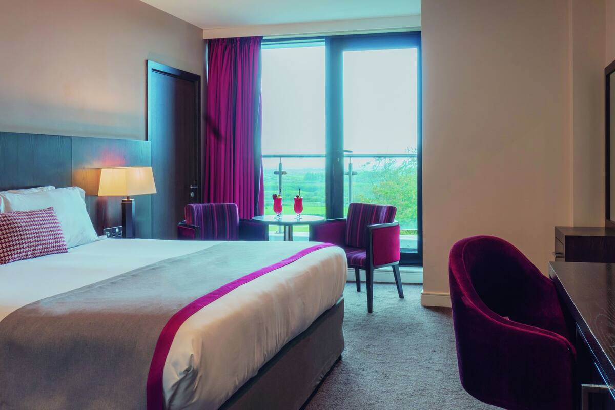 A hotel room with a large bed, a desk, a chair, and a balcony with a view.