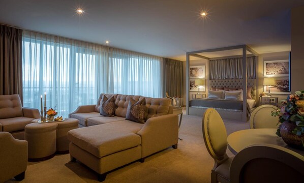 A spacious living room with a sofa, ottoman, and a dining area with a chair. There is a bedroom with a bed and a nightstand.