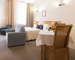 A hotel room with a bed, two armchairs, a dining table with a vase of flowers, and a view of the outdoors.