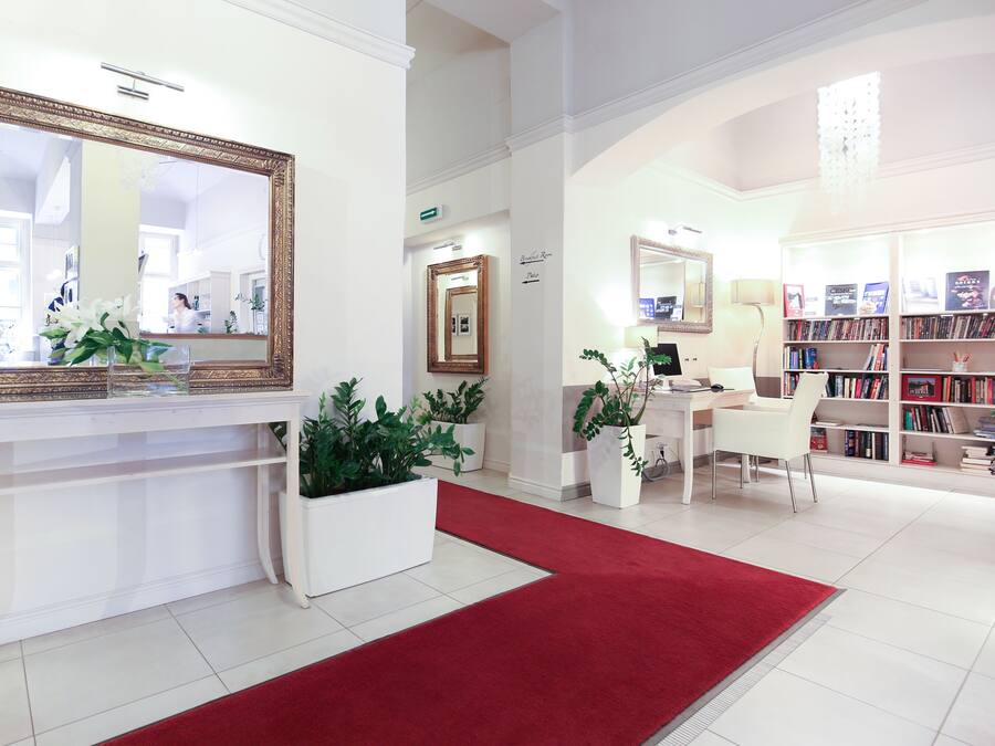 A hotel lobby with a reception desk, a red carpet, potted plants, a desk with a computer, a chair, and a bookshelf filled with books.
