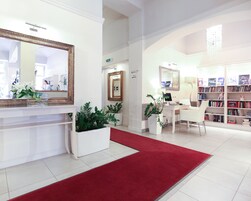 Vestíbulo de hotel con mostrador de recepción, alfombra roja, plantas en macetas, escritorio con computadora, una silla y una estantería llena de libros.