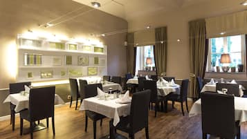 Interior de restaurante con varias mesas y sillas, una pared decorada con cuadros, y ventanas amplias con cortinas.