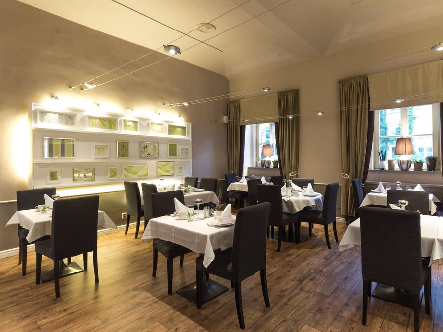 Interior de restaurante con varias mesas y sillas, una pared decorada con cuadros, y ventanas amplias con cortinas.