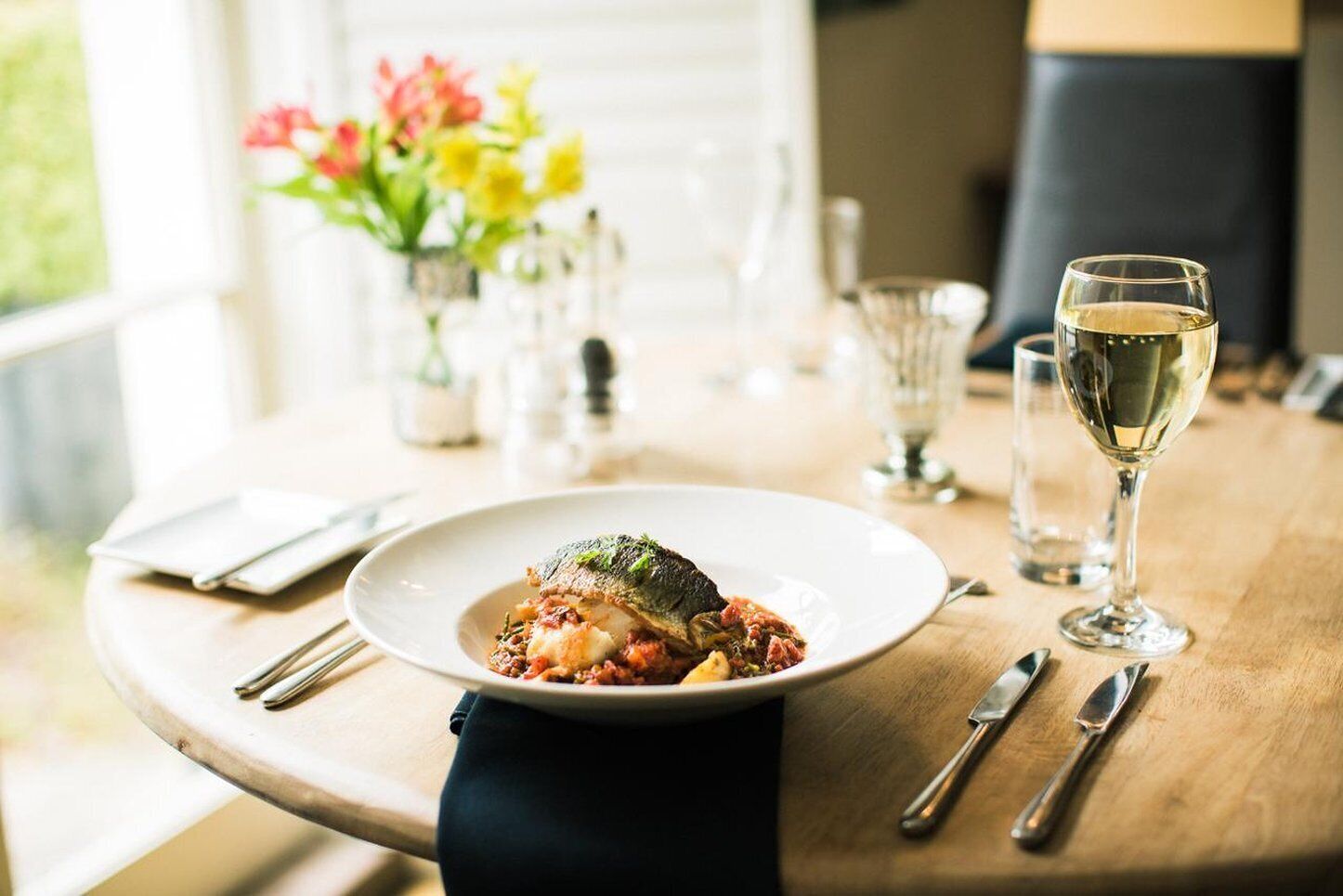 A dining table with a plate of food, a glass of white wine, and cutlery.