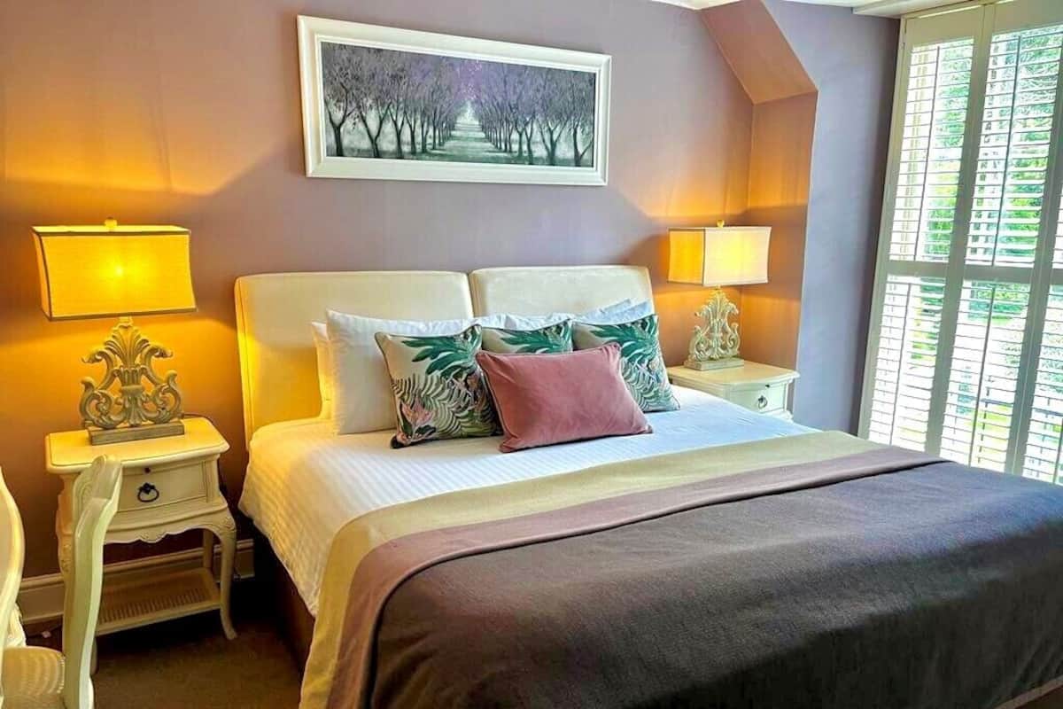 A bedroom with a large bed, two bedside tables, a nightstand, a framed picture on the wall, and a window with blinds.