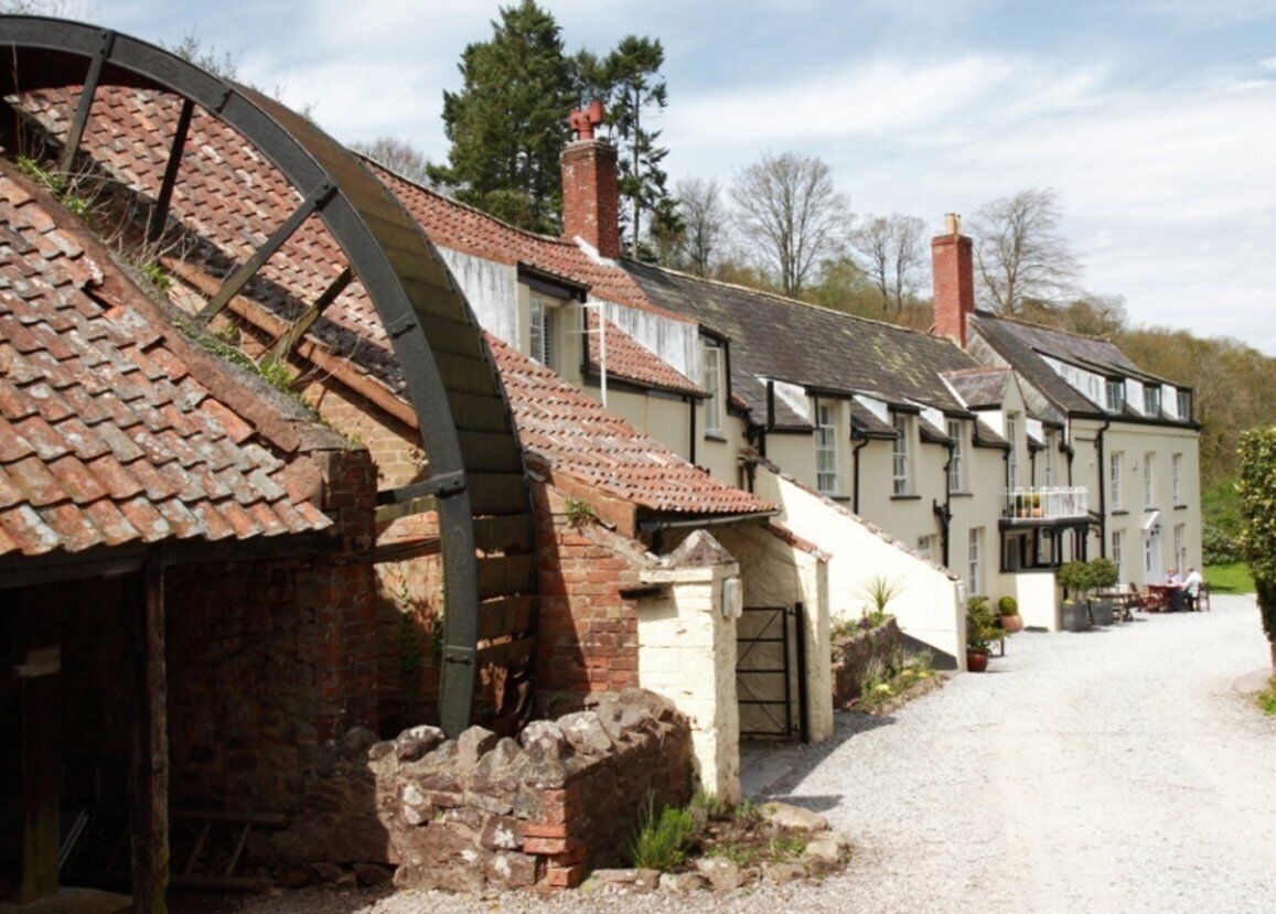 A traditional building with a large water wheel, a gravel driveway, and a small garden area with a table and chairs.
