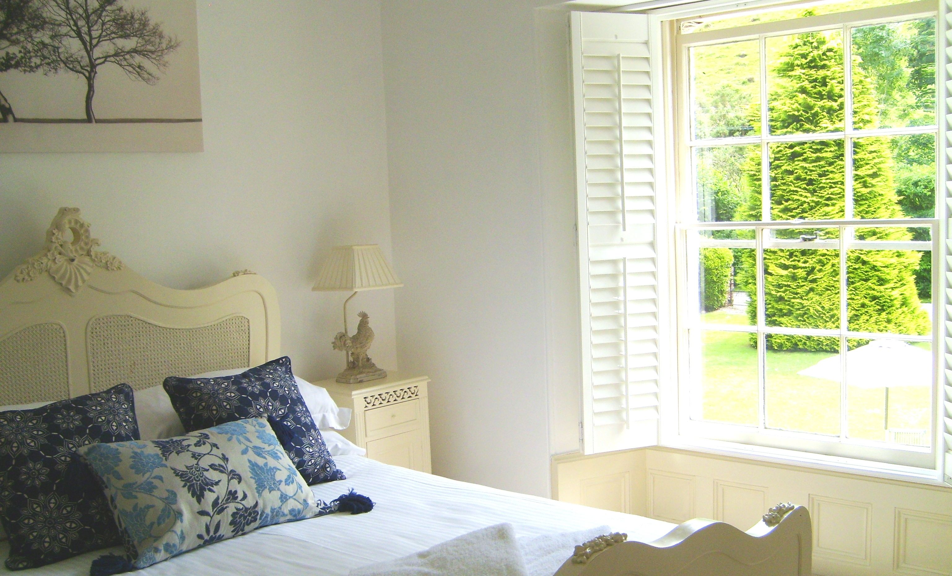 A bedroom with a bed, pillows, a nightstand, a lamp, and a window with a view of greenery.
