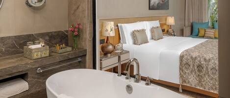 A modern hotel room with a large bathtub, a separate bedroom with a bed, a vanity with a mirror, and a window with curtains.