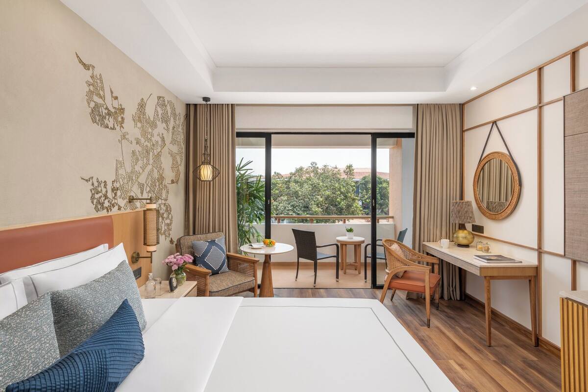Habitación de hotel con cama, escritorio, silla, mesita y balcón con vistas.