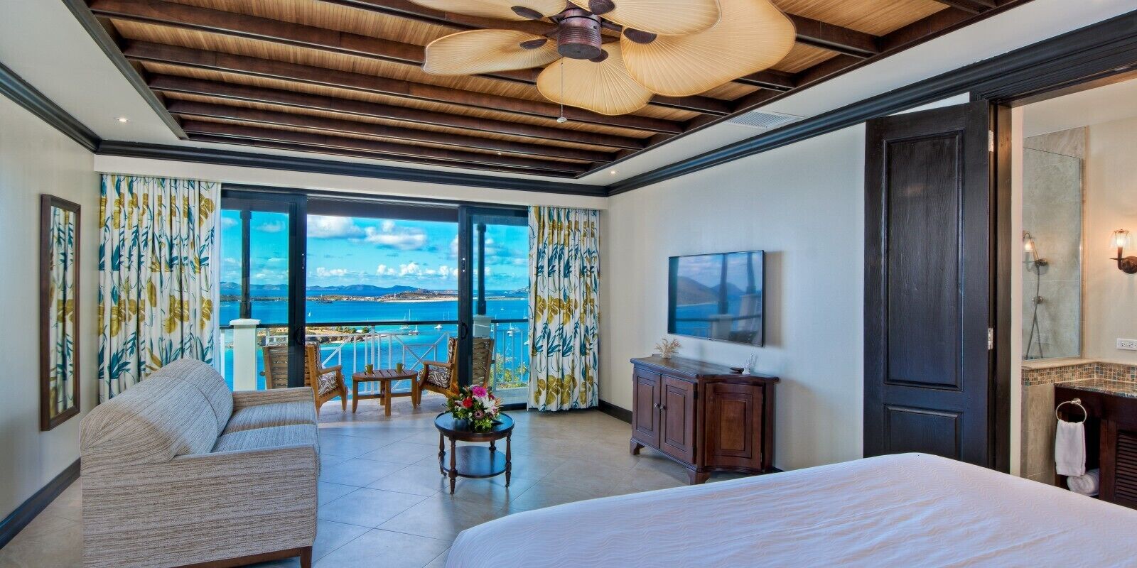 Habitación de hotel con una cama grande, zona de estar con sofá y sillones, una mesita con flores, ventilador de techo y vista al mar a través de amplias ventanas.
