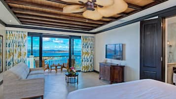 Habitación de hotel con una cama grande, zona de estar con sofá y sillones, una mesita con flores, ventilador de techo y vista al mar a través de amplias ventanas.