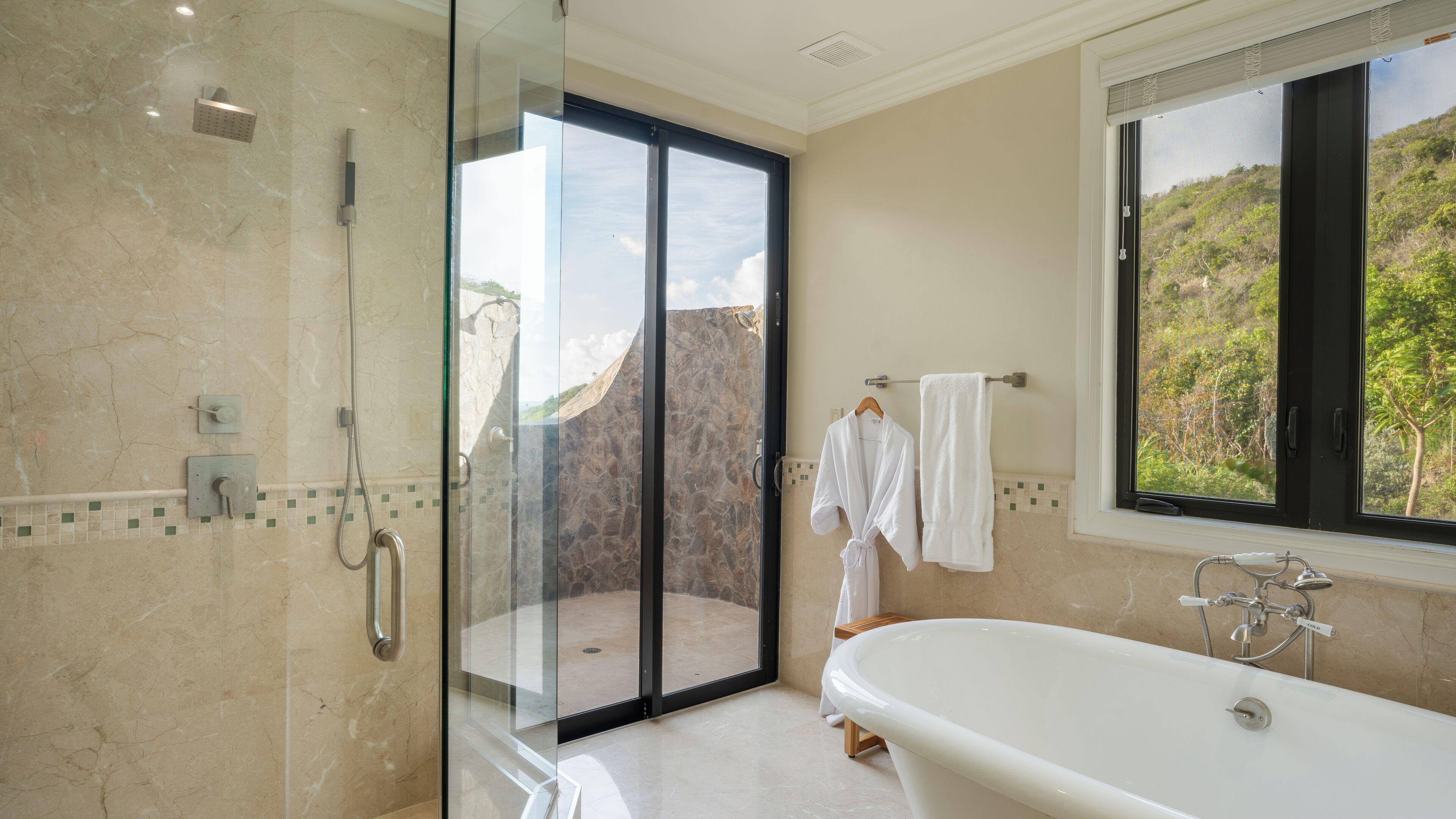 A bathroom with a glass-enclosed shower, a bathtub, and a window with a view of greenery.