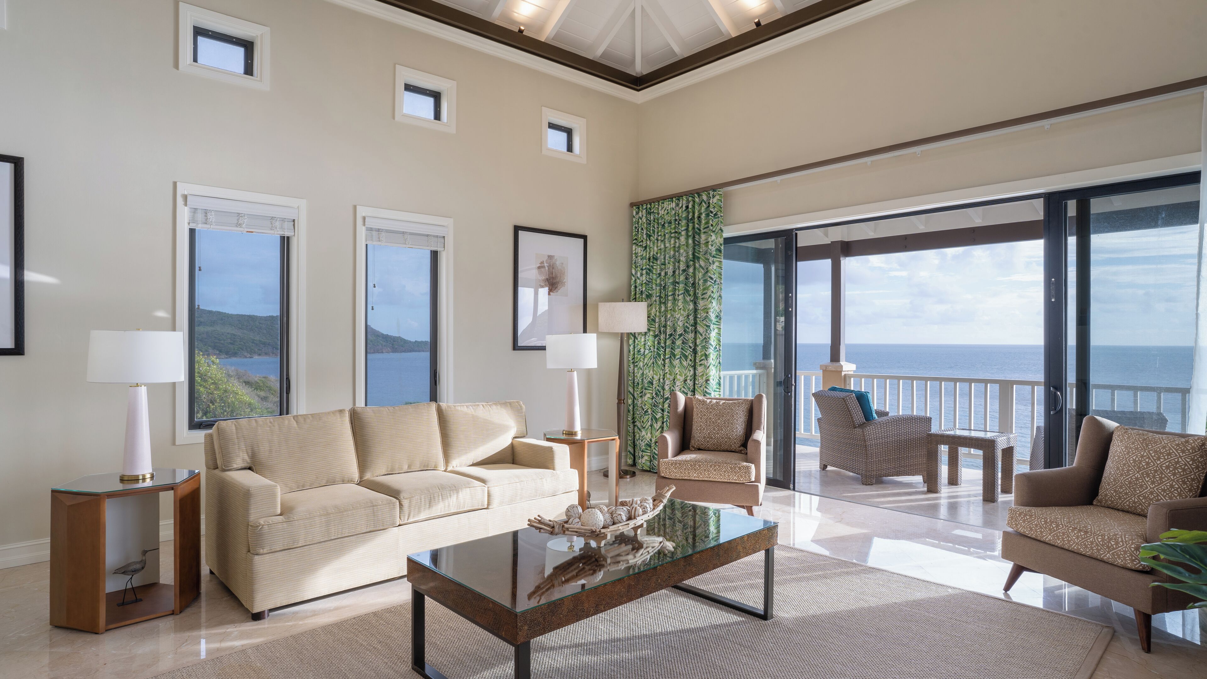 Un ampio soggiorno con una grande vetrata che dà su un balcone con vista mare. Si può vedere un divano color beige, un tavolino in vetro e due poltrone.