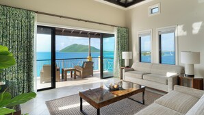 A spacious living room with a large sliding glass door leading to a balcony with a view of the sea and a mountain. There is a sofa, a coffee table, and a lamp. - Scrub Island Resort, Spa & Marina (Scrub Island)