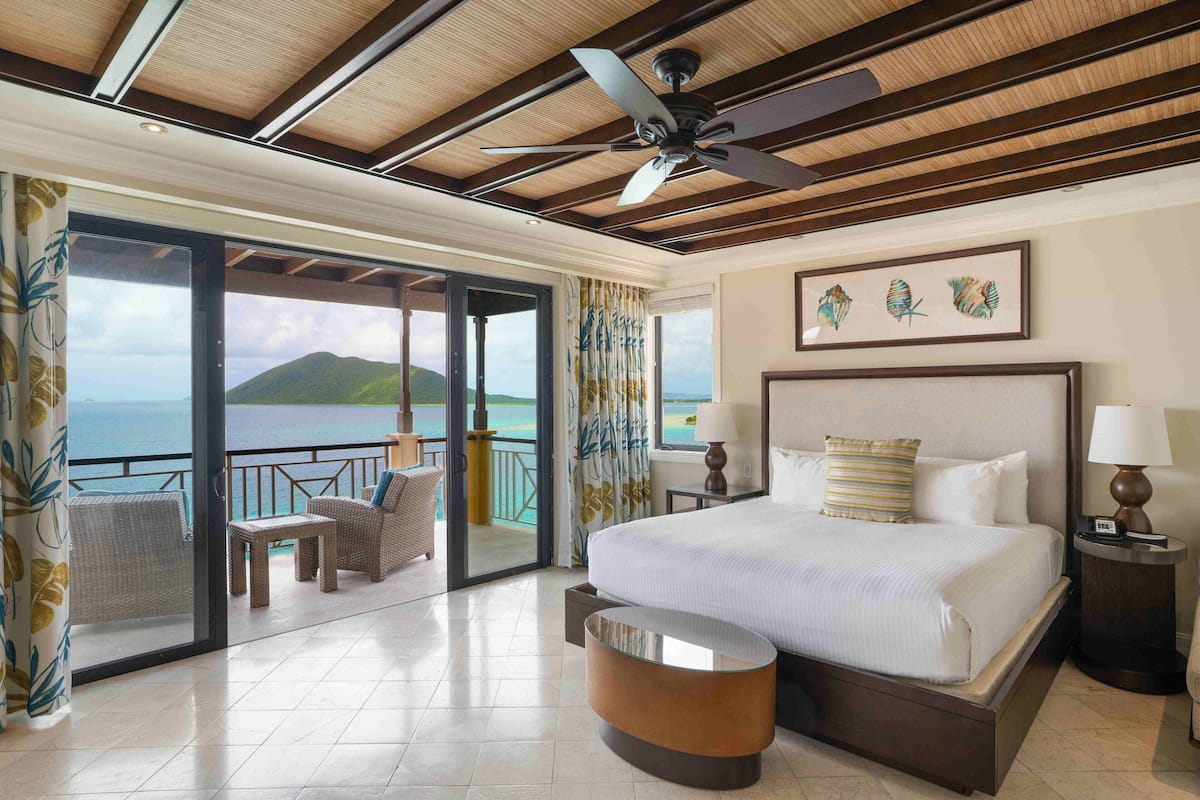 Habitación de hotel con una cama grande, un ventilador de techo, un balcón con vistas al mar y una zona de comedor con muebles de mimbre.