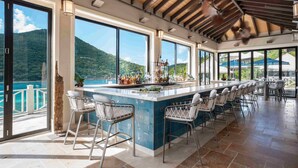 A bar area with a long counter, multiple bar stools, and large windows offering a view of the sea and mountains. - Scrub Island Resort, Spa & Marina (Scrub Island)