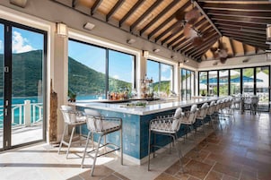 Zona de bar con una barra alargada, varios taburetes y amplias ventanas con vistas al mar y a las montañas.