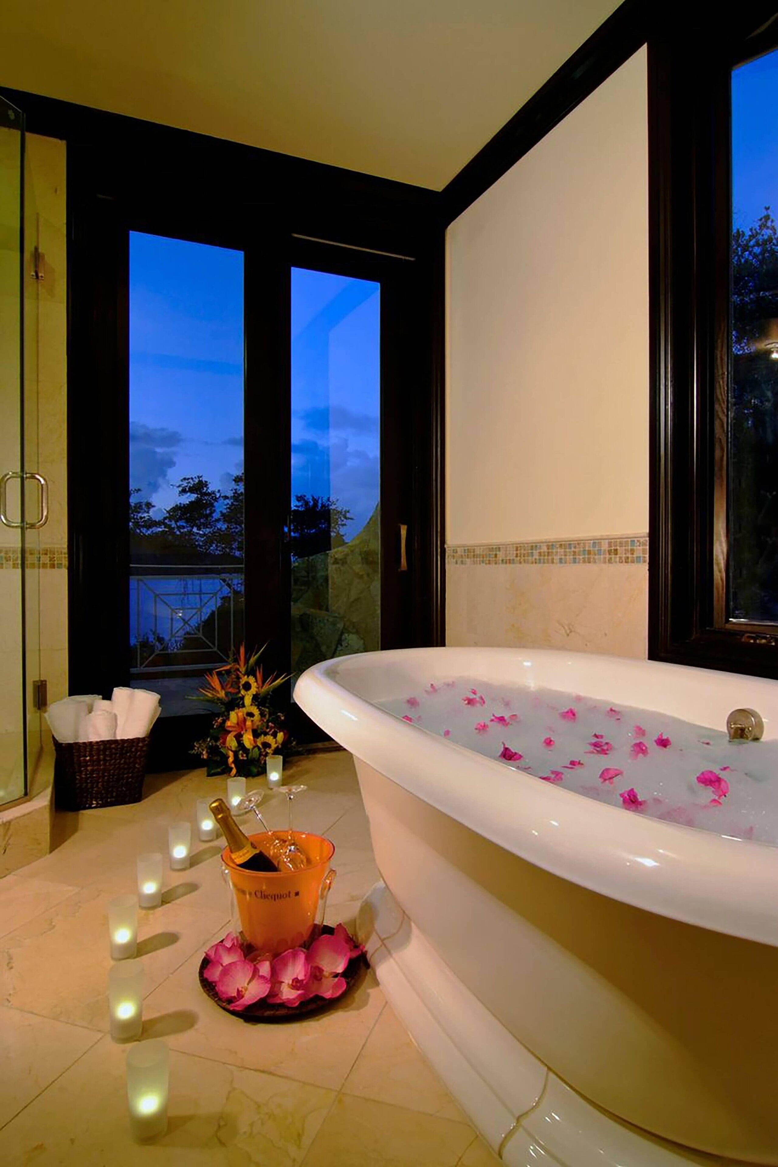 A bathroom with a large bathtub filled with rose petals, a champagne bottle, and a flower arrangement.