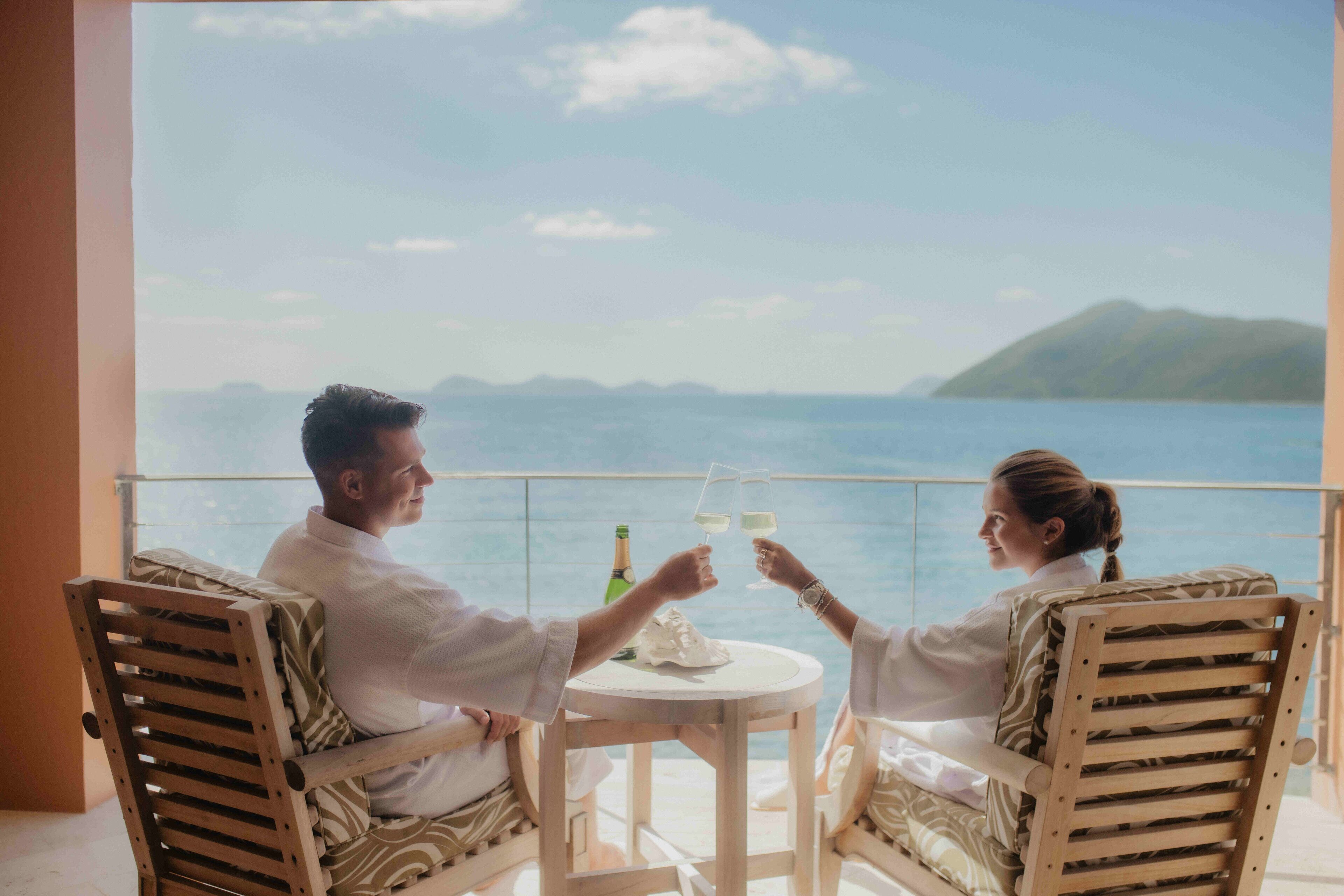 Un couple est assis à une table ronde, devant une bouteille et des verres, avec une vue sur une étendue d’eau et un paysage lointain.