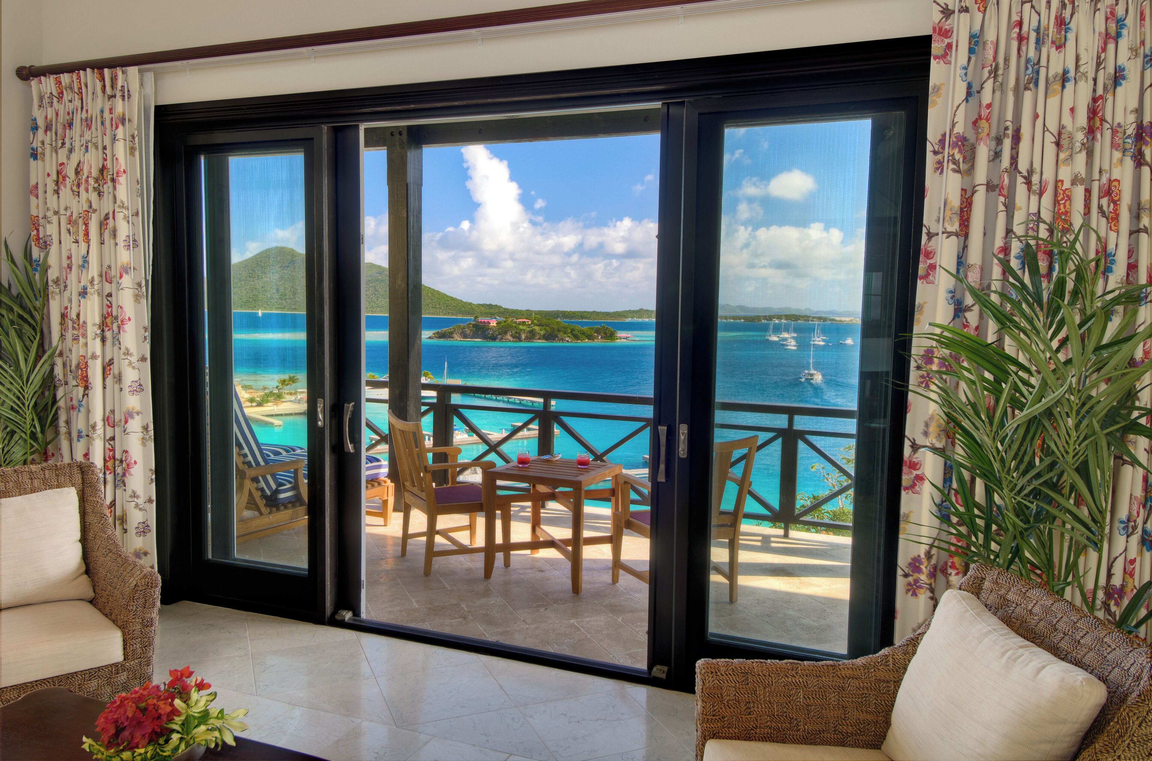 Habitación con puerta de vidrio corrediza que da a un balcón con vistas al mar, una piscina y una pequeña isla. En el balcón hay sillas de mimbre y una mesa.