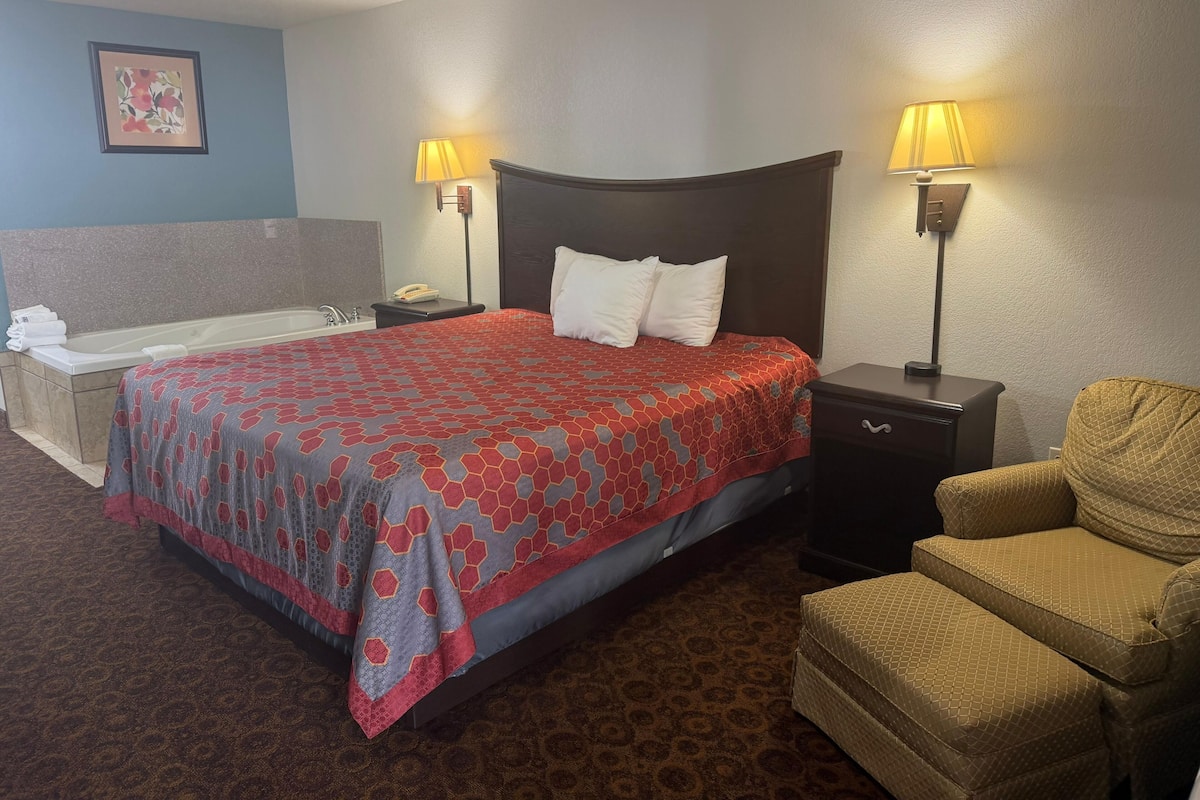 A hotel room with a bed, two bedside lamps, a chair, a small table, a nightstand, a bathroom with a bathtub, and a towel rack.