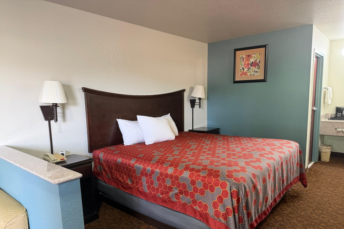 A hotel room with a bed, two bedside lamps, a nightstand, a small table, a wall-mounted picture, and a bathroom door.
