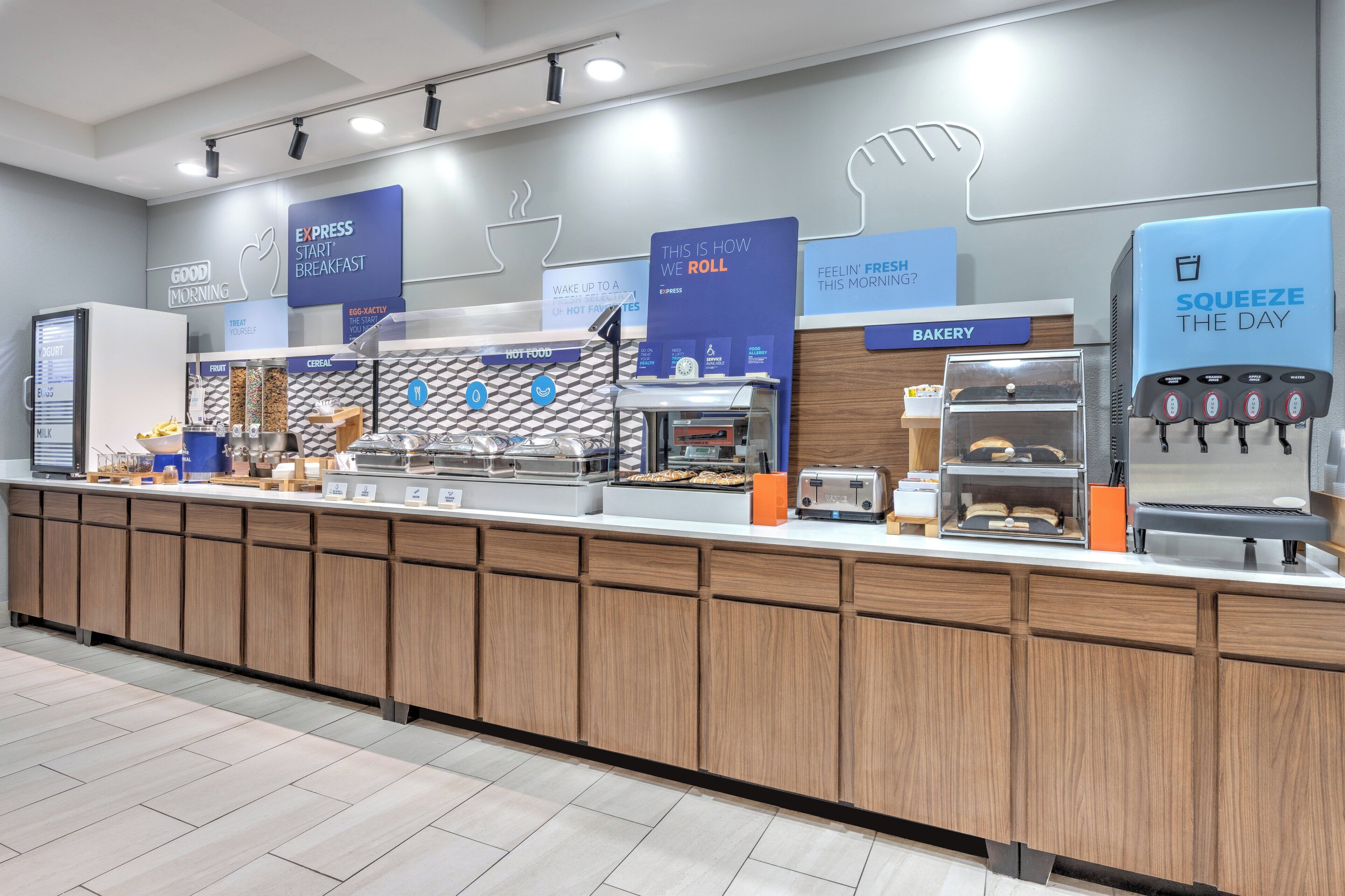 A modern breakfast area with a counter displaying various food items, a coffee machine, and a sign indicating the bakery section.
