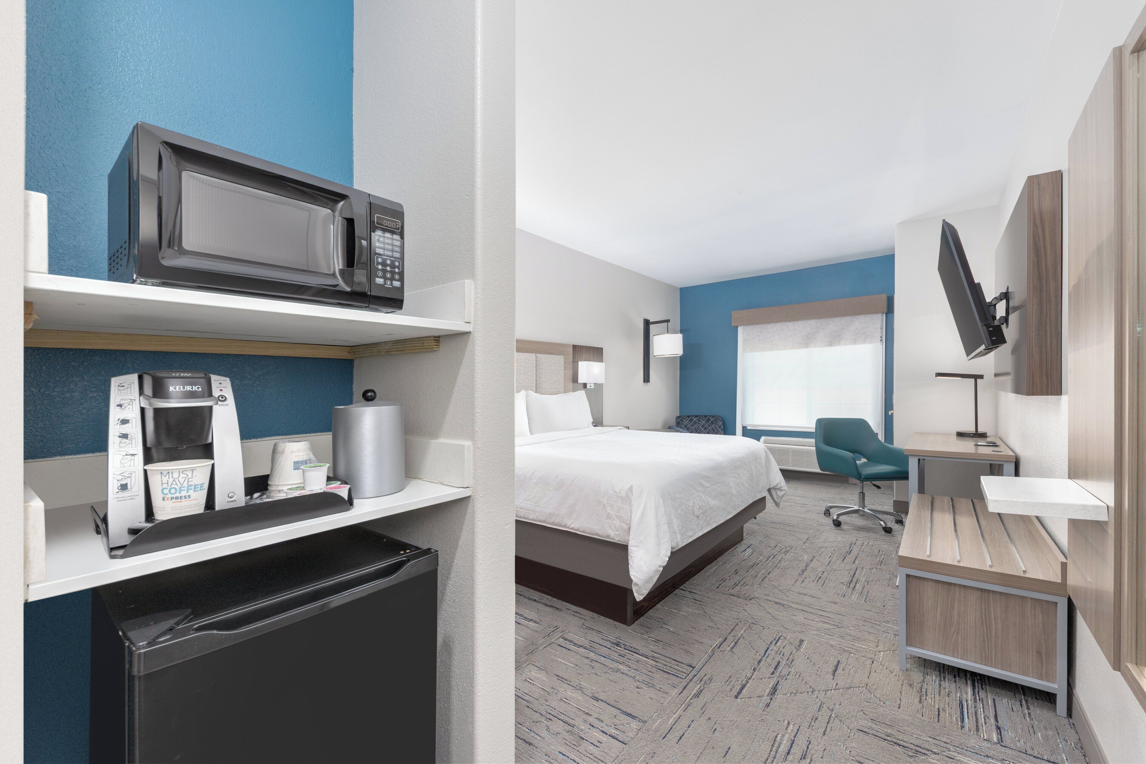 A hotel room with a bed, a desk, a chair, a microwave, a coffee machine, and a television.