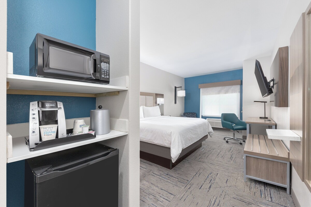 A hotel room with a bed, a desk, a chair, a microwave, a coffee machine, and a television.