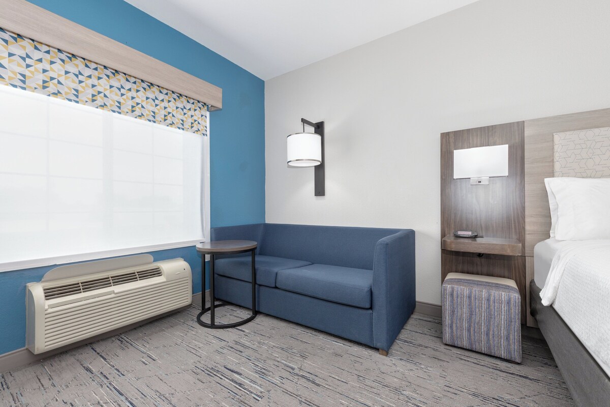 A hotel room with a blue wall, a white air conditioning unit, a blue sofa, a small round table, a bedside table with a lamp, and a bed with a white comforter.