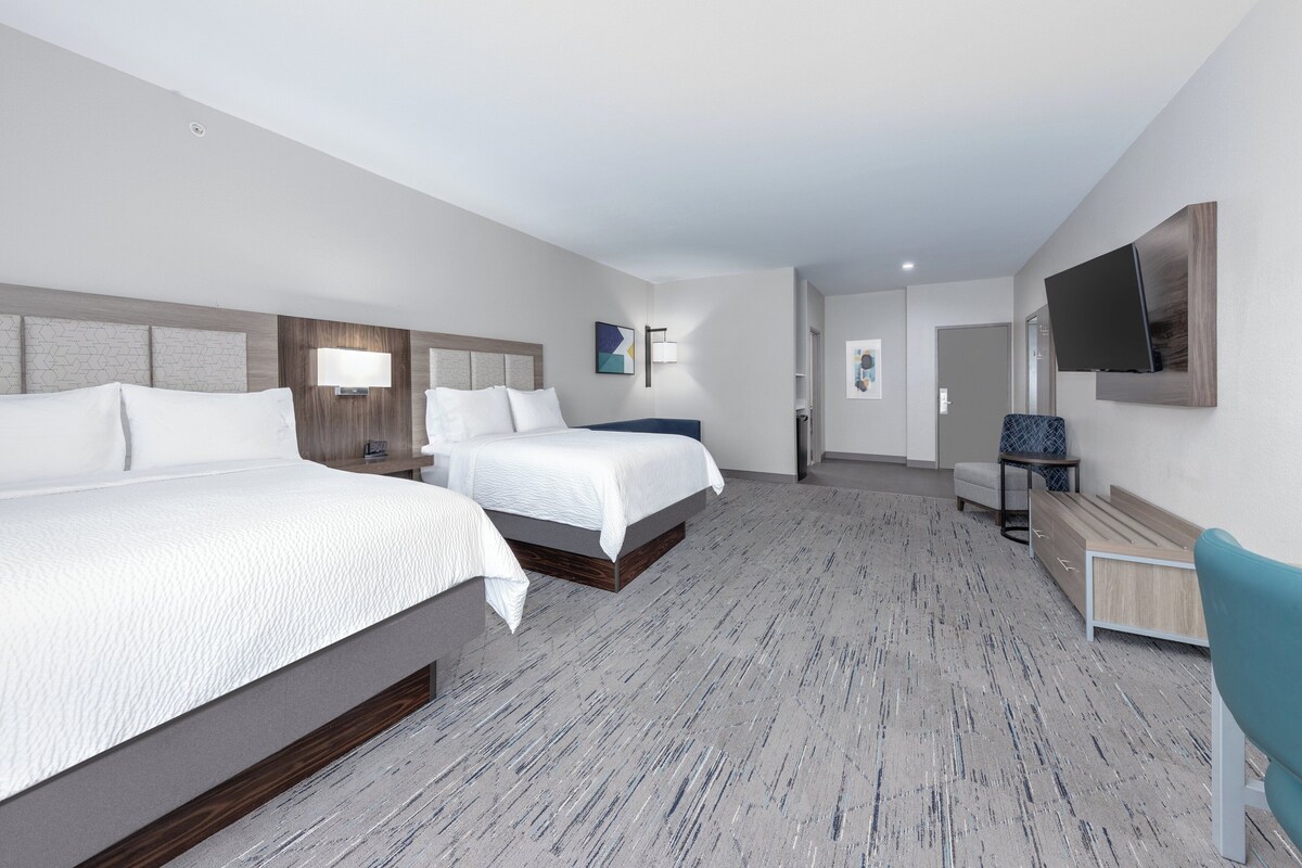 A hotel room with two beds, a desk, a chair, a flat-screen TV, and a large window.