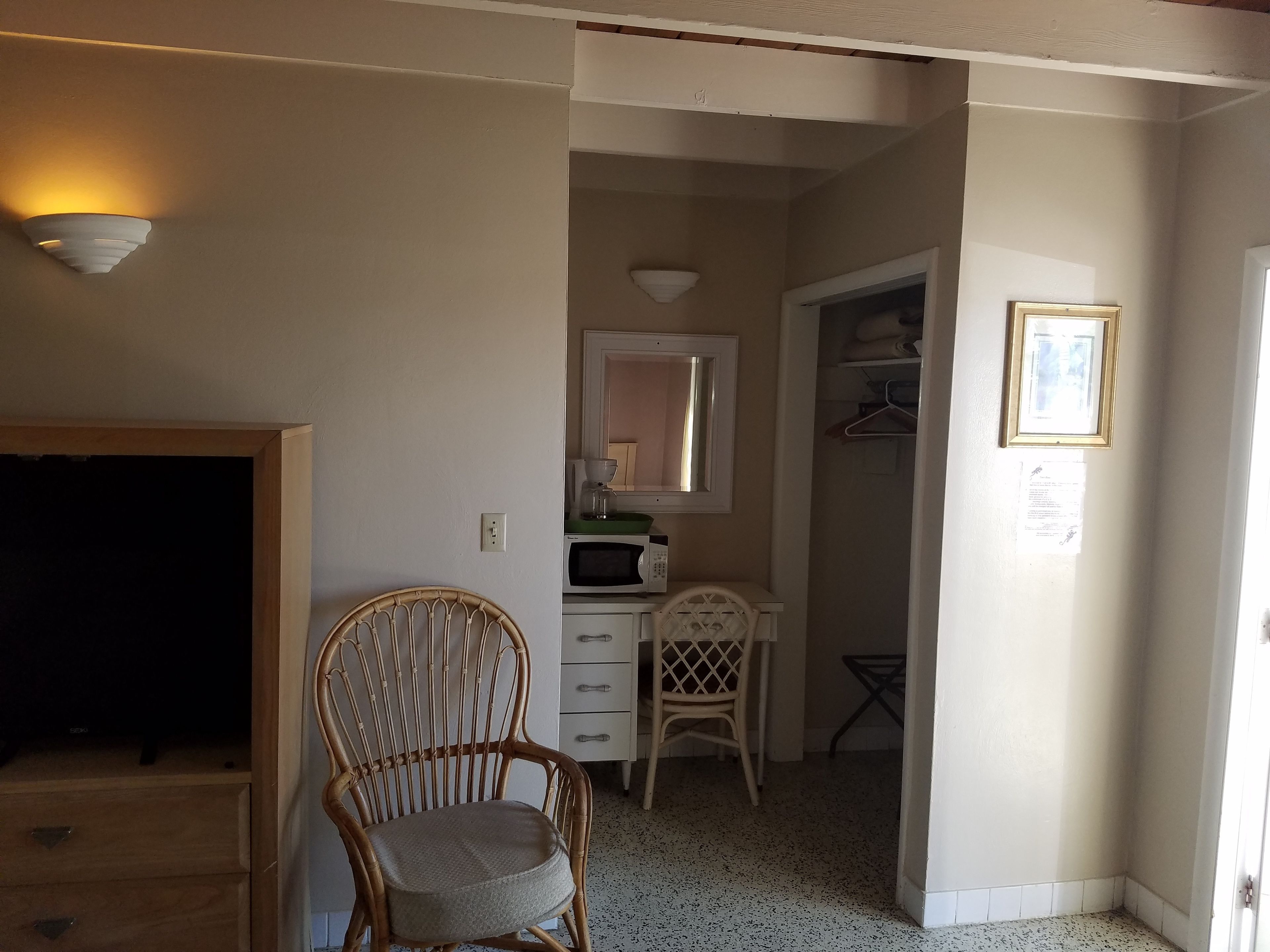 Una habitación con una silla de mimbre, un escritorio con espejo, un microondas y una mesita.