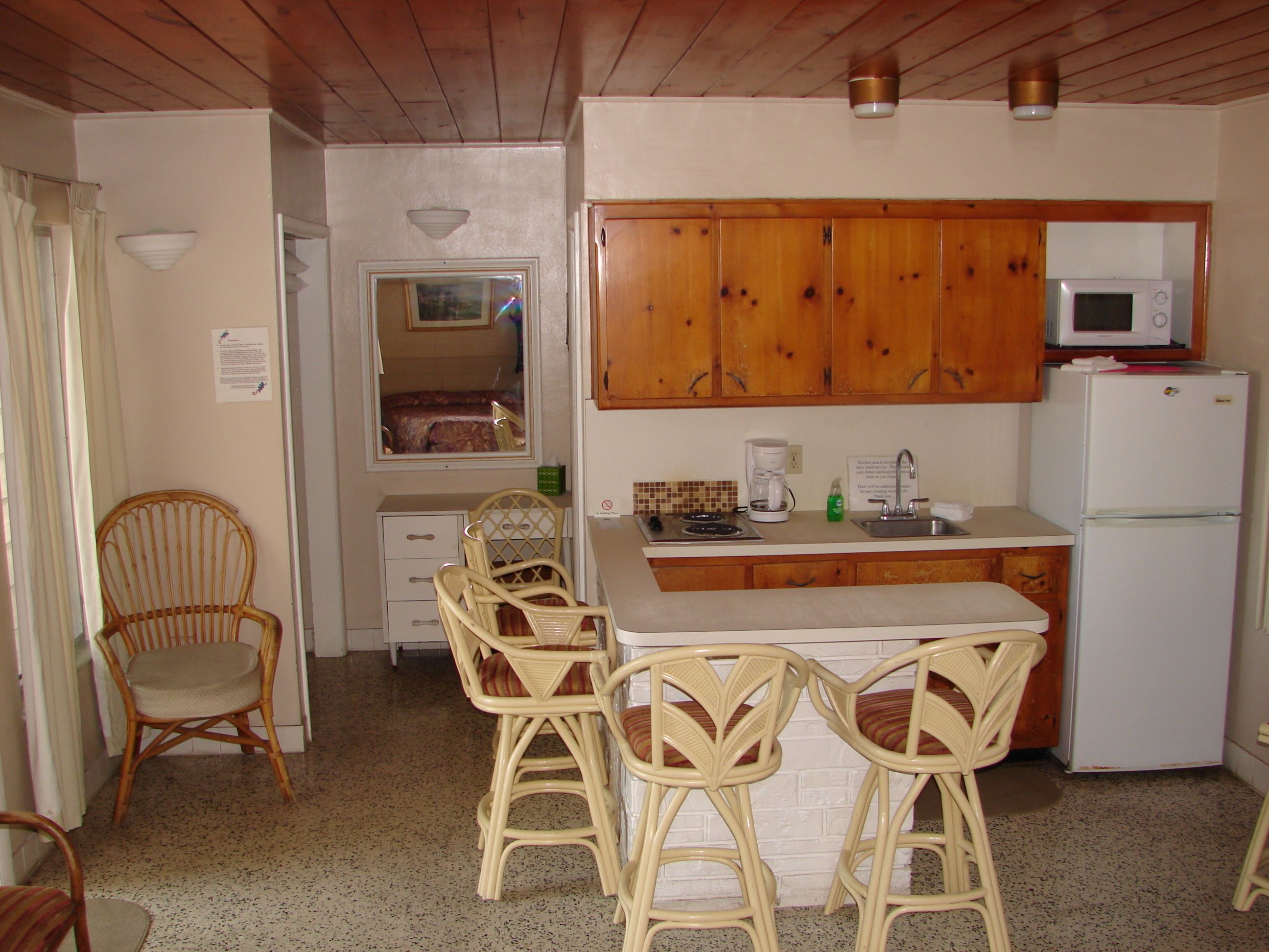 Una cocina compacta con armarios de madera, un microondas, un refrigerador y un pequeño comedor con sillas de mimbre.