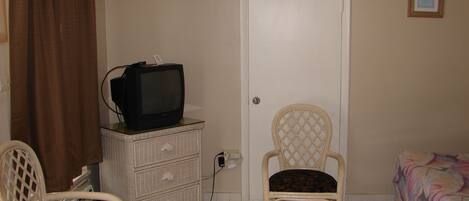 A room with a television on a dresser, a chair, a bed, and a door.