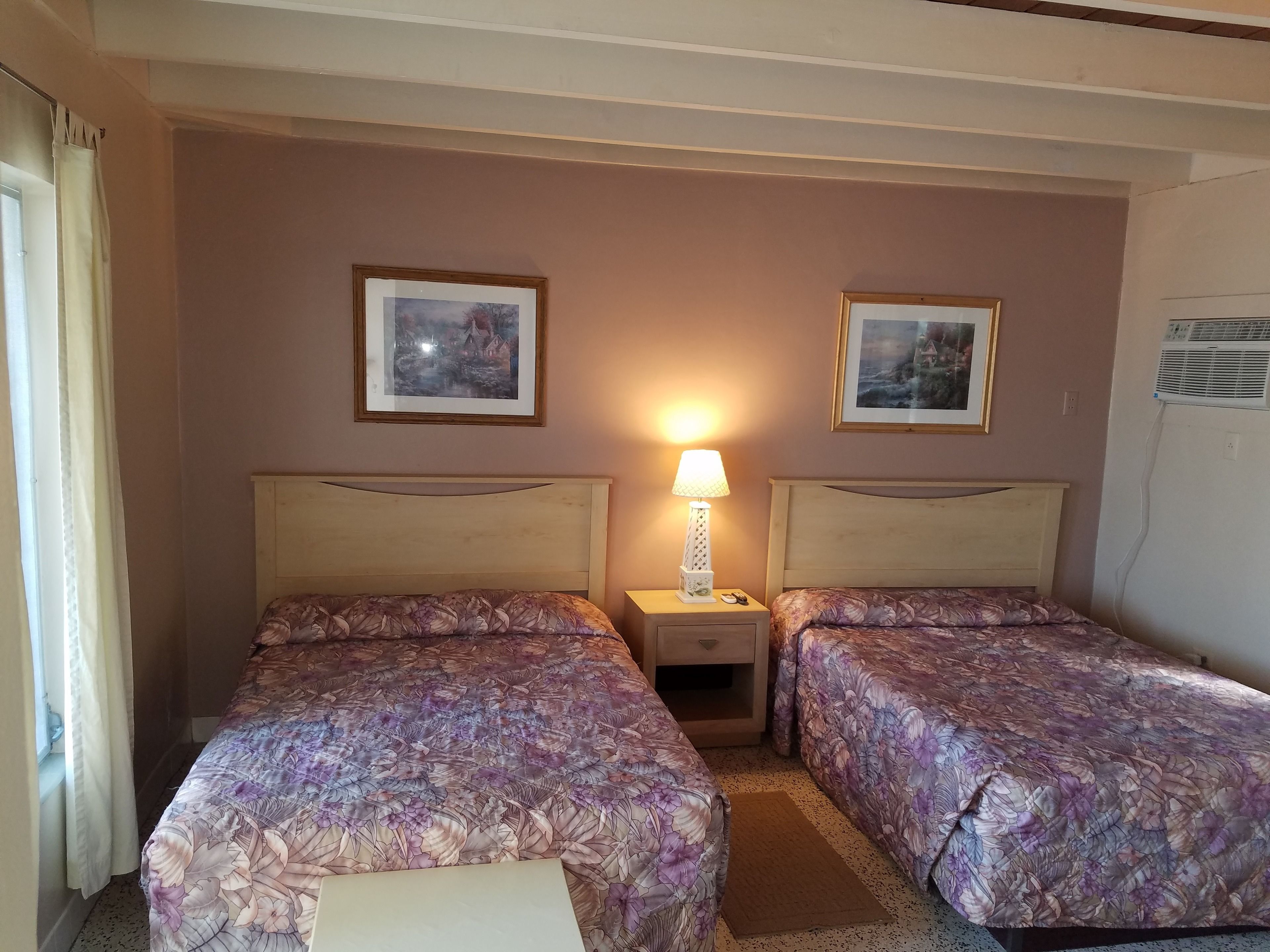 Una habitación de hotel con dos camas, una mesita de noche con lámpara y dos cuadros en la pared.