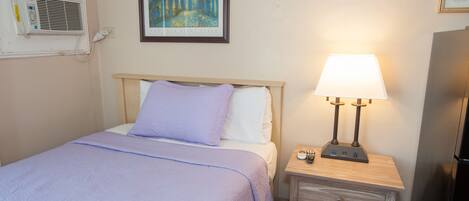 A hotel room with a bed, a nightstand with a lamp, a painting on the wall, and a refrigerator.