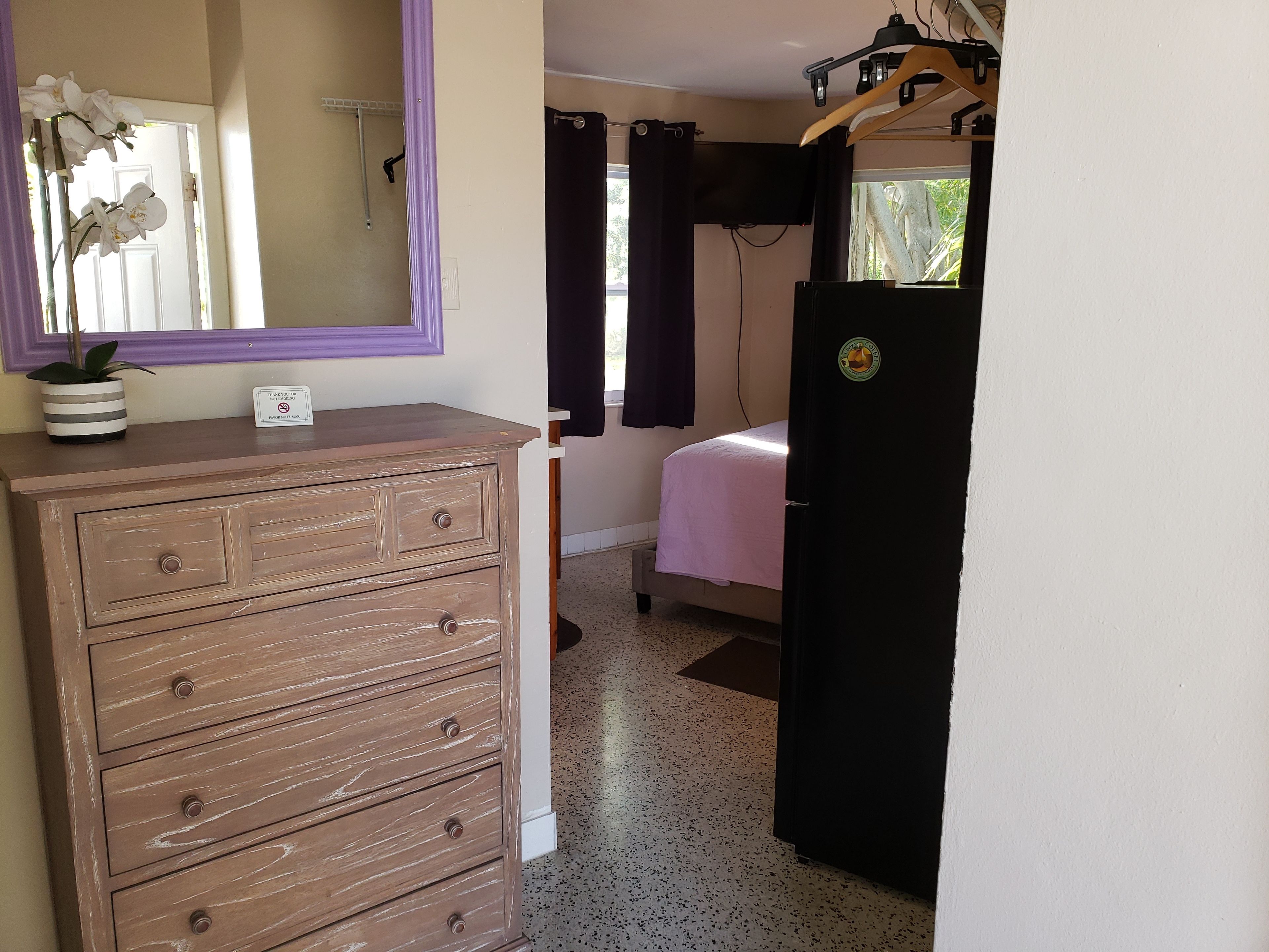 A bedroom with a wooden dresser, a bed with a pink bedspread, a window with curtains, and a door with a sticker.