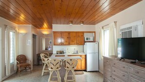 Cocina compacta con armarios de madera, microondas y refrigerador. Hay una zona de comedor con sillas de mimbre y una mesa pequeña.