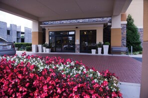 A hotel entrance with a paved walkway, a car parked in front, and a flower bed with red and white flowers. - Holiday Inn Express & Suites Atlanta East – Lithonia by IHG (Lithonia)